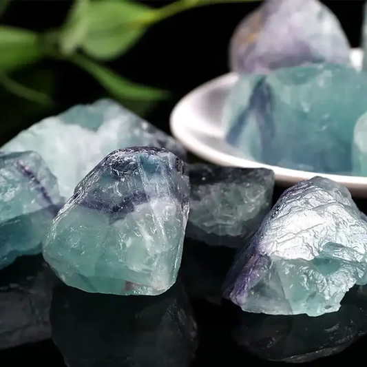 Rough Fluorite Crystal stones in light blue, purple, and grey tones. The raw fluorite stones are displayed on a reflective surface with a bowl in the background.